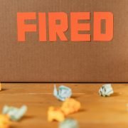 Close-up of a cardboard box labeled FIRED on a wooden table with crumpled papers.