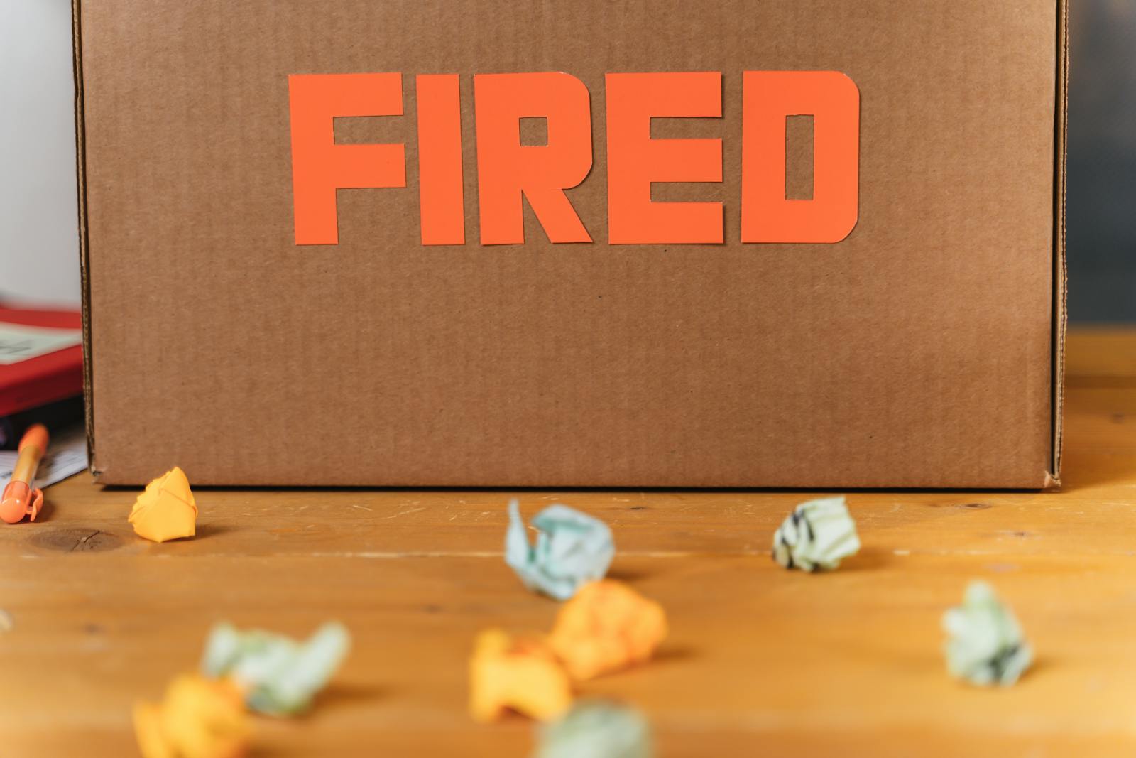 Close-up of a cardboard box labeled FIRED on a wooden table with crumpled papers.